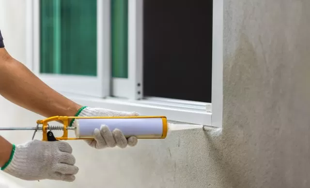 A homeowner carefully applying caulk to the window sill of their house, creating a secure, weather-resistant seal.